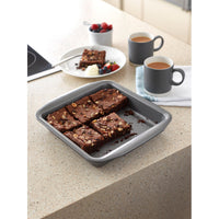 Square non-stick cake tin with chocolate brownies sat on table with mugs of tea.