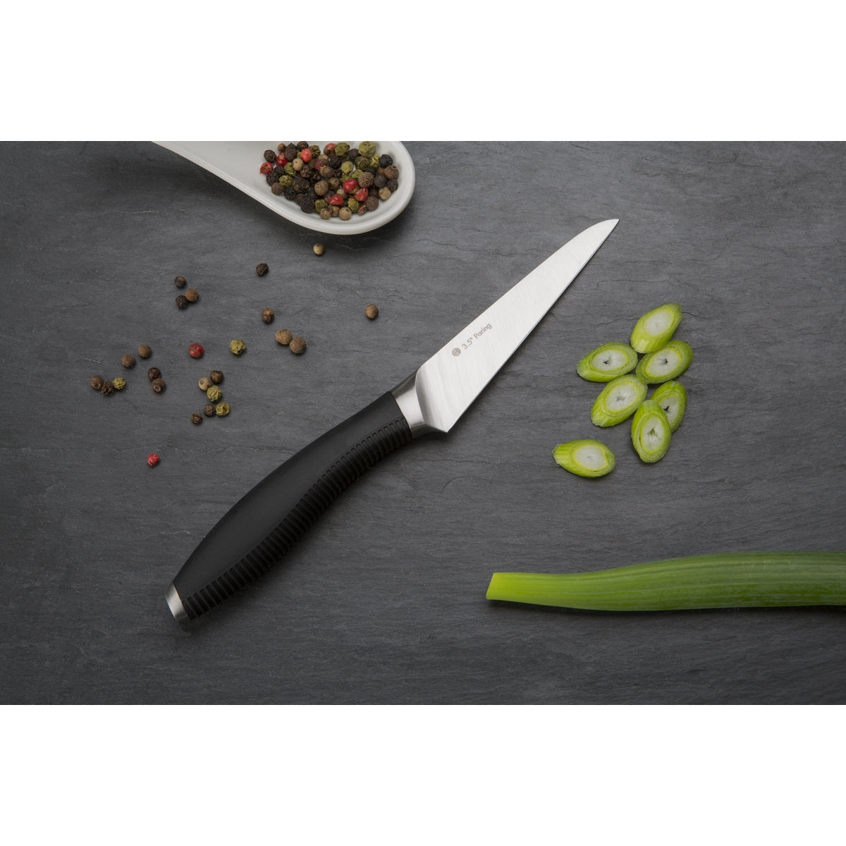 Image shows small steel paring knife on a grey background with sliced spring onion and peppercorns.